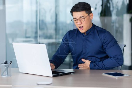 Téléchargez les photos : Sick office worker at workplace with laptop inside office, asian man got food poisoning and has severe stomach pain, businessman in shirt and glasses holds hand on stomach grimaced in pain. - en image libre de droit