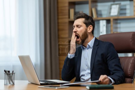 Foto de Mature man yawning inside classic office, senior businessman in workplace sleepless and overworked, boss in business suit sitting at desk, using laptop at work. - Imagen libre de derechos