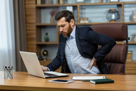 Téléchargez les photos : Mature man working in modern office with laptop, senior boss having severe back pain, businessman at workplace overworked working late massaging his back with hand, severe pain from sitting. - en image libre de droit