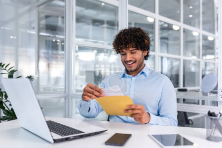 Young successful businessman inside office working with laptop, man received good news notification mail, hispanic man reading news and smiling at workplace, happy with achievement result.