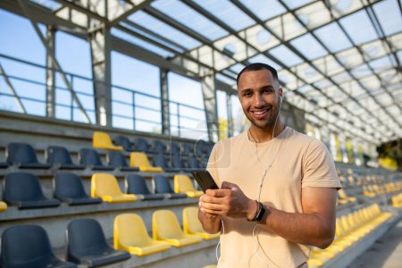 Photo for Young African American male athlete, coach, runner wearing headphones stands in stadium between bleachers, uses mobile phone. He looks at the camera, smiles. - Royalty Free Image