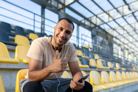 Photo for Portrait of a young African American male athlete, coach sitting in a stadium wearing headphones and using the phone. He points at the camera with a super finger, smiles, rests. - Royalty Free Image