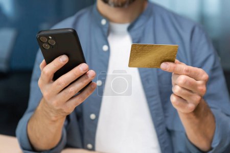 Photo for Close-up of mans hands inside the office, businessman in blue shirt and white t-shirt works at workplace, man holds bank credit card and smartphone, makes online purchase in online store. - Royalty Free Image