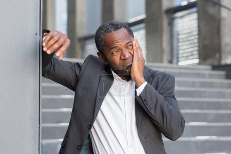 Toothache on the street. A senior African American man in a suit stands outside an office and holds his cheek, suffering from a toothache.