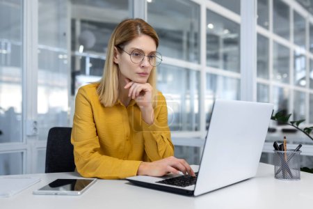 Portrait of a serious thinking businesswoman inside a bright office at work with a laptop, a woman in a yellow shirt and glasses works thoughtfully while sitting at a workplace