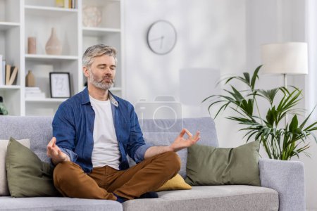 Photo for Adult mature man alone at home relaxing sitting in lotus position on sofa, gray haired person relaxing meditating indoors in living room in casual clothes. - Royalty Free Image