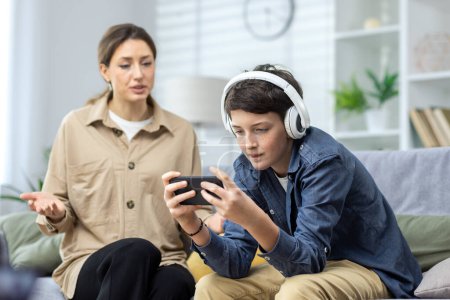 Photo for Family conflict quarrel, woman mother quarrels with son, teenage boy in headphones ignores woman and plays video games on phone, family at home in living room sitting on sofa. - Royalty Free Image