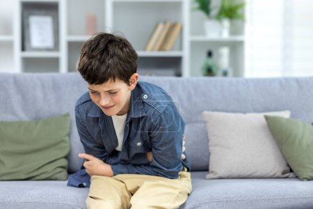 Foto de Un niño, un niño, un adolescente, se sienta solo en casa en el sofá y sostiene su estómago. Sufrir de dolor, agacharse, necesita ayuda médica. - Imagen libre de derechos