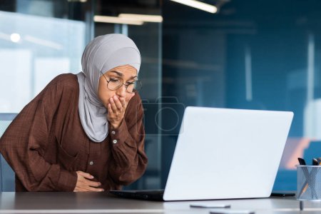 Téléchargez les photos : Femme d'affaires malade dans le hijab travaillant à l'intérieur du bureau sur le lieu de travail, nausées et douleurs d'estomac chez les femmes musulmanes, femme vomissant de la nourriture, vomissant assis à table avec un ordinateur portable. - en image libre de droit