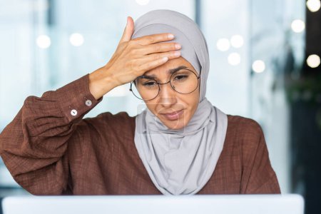 Photo for Tired business woman working at workplace inside office, Muslim woman with hijab is sick with severe headache, female worker uses laptop at work. - Royalty Free Image