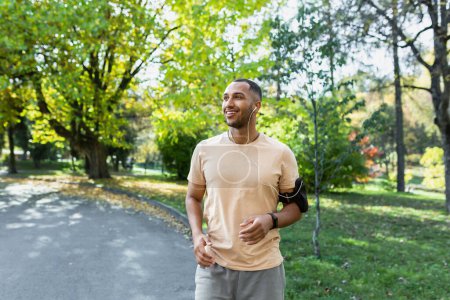 Photo for Morning run. Healthy Lifestyle. Young hispanic man running in the park wearing headphones and holding a phone. - Royalty Free Image