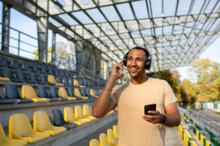 Photo for Morning jogging, exercises. Healthy Lifestyle. Young hispanic man doing sports in the stadium, standing among the bleachers wearing headphones and holding a phone. - Royalty Free Image