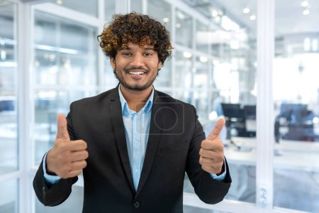 Photo for Portrait of successful hispanic businessman boss, manager in business suit and beard looking at camera and smiling standing near window, showing thumbs up, sign of success and achieving goals. - Royalty Free Image