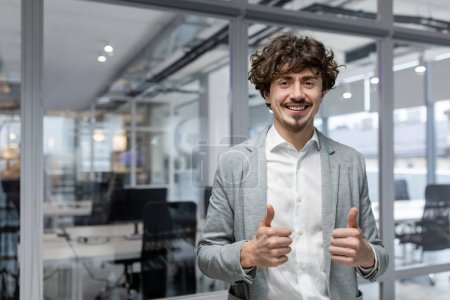 Photo for Portrait of successful businessman boss, manager in business suit and beard looking at camera and smiling standing near window, showing thumbs up, sign of success and achieving goals - Royalty Free Image