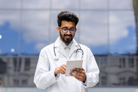 Young successful Hindu student in white medical coat walks outside clinic, doctor intern trainee with tablet computer in hands, browsing online educational material.