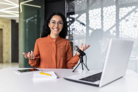 Portrait of young Hispanic woman sitting in office at desk in front of microphone and laptop and recording blog, podcast, webinar. Smiling at the camera.
