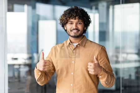 Photo for Portrait of successful hispanic businessman boss, manager looking at camera and smiling standing near window, showing thumbs up, sign of success and achieving goals. - Royalty Free Image