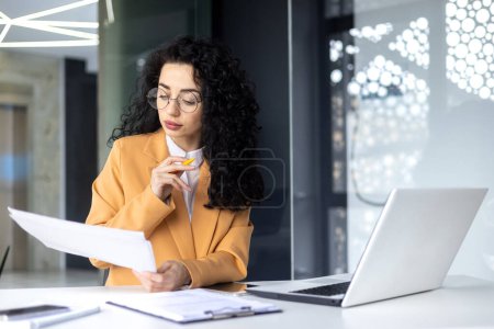 Photo for Serious focused female financier working inside office with documents and papers at workplace, hispanic woman thinking about bills on paper work. - Royalty Free Image