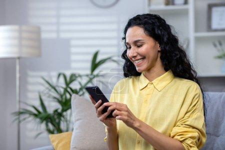 Photo for Young beautiful Latin American woman using phone alone at home, woman smiling and happy sitting on sofa in living room browsing online sites and using app on smartphone. - Royalty Free Image