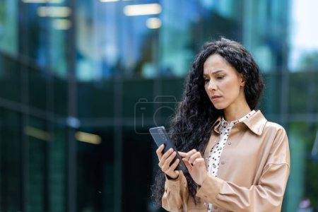 Photo for Upset and sad woman with phone in hands reading online message, Hispanic business woman received bad news notification while walking outside office building with smartphone. - Royalty Free Image