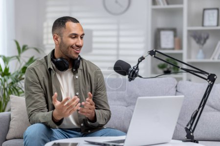 Junger hispanischer Mann sitzt in einem kleinen Heimstudio. Sitzt mit Kopfhörern auf dem Sofa und nimmt eine Sendung auf, kommuniziert über ein Mikrofon über einen Video-Online-Laptop.