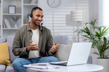 Young Latin American businessman, freelancer works remotely from home. Sitting on the sofa in a headset and talking on a video call through a laptop.
