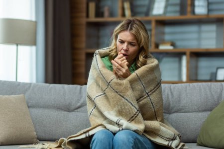 Single mature woman sitting at home in the living room on the sofa, wrapped in a blanket trying to keep warm, missing and broken heating in the house.