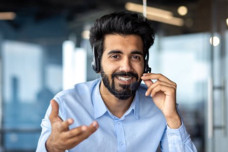 Photo for Close-up photo. Portrait of a young Indian man in a headset sitting in front of the camera, smiling, talking, consulting, gesturing with his hands. - Royalty Free Image