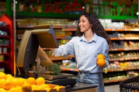 Foto de Mujer joven compradora en el supermercado utiliza balanzas de autoservicio, la sonriente mujer hispana pesa frutas y verduras en una gran tienda. - Imagen libre de derechos