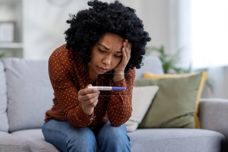 Photo for Frustrated and sad young african american woman crying sitting on sofa at home and holding negative pregnancy test. - Royalty Free Image