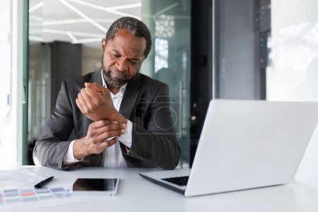Overtired and overworked man at workplace sitting inside office, businessman has wrist joint pain on his hand, african american man in business suit working long time sitting.