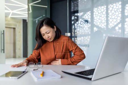 Photo for Sick woman at workplace has acute stomach pain, asian business woman holding hand on stomach from pain, asian woman working sitting at desk with laptop inside office. - Royalty Free Image
