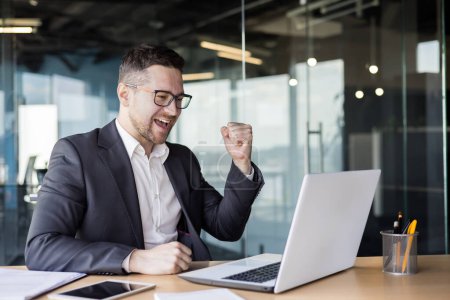 Photo for Happy young male businessman sitting in front of laptop in office and enjoying success, showing victory gesture with hand. - Royalty Free Image