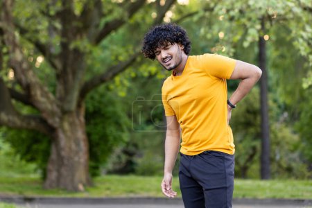 Photo for A young Indian man injured and sprained his back while jogging in a park outside. He stands holding his hand to his body and grimaces in pain. - Royalty Free Image