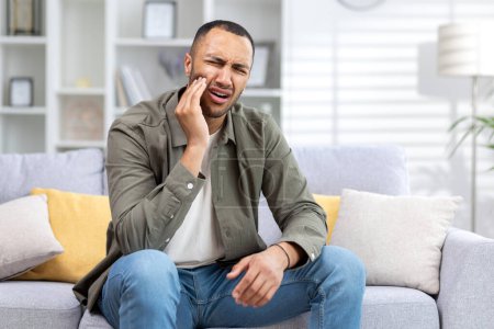 Téléchargez les photos : Jeune homme hispanique souffrant de maux de dents assis sur le canapé à la maison et tenant la main à la joue. - en image libre de droit