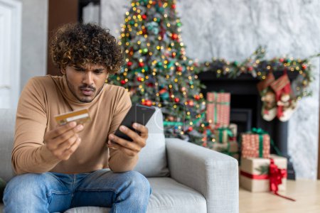 triché et bouleversé homme de Noël dans le salon près décoré arbre de Noël, rejeté et mauvais transfert d'argent, homme hispanique tenant carte de crédit bancaire et téléphone.