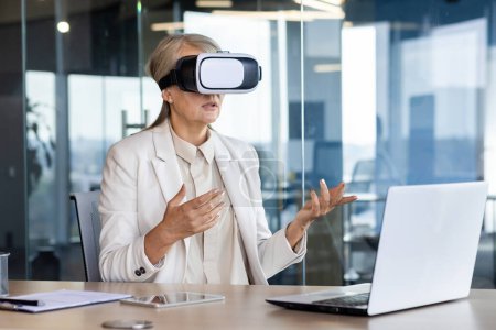 Senior experienced business woman using virtual reality glasses for remote meeting, video call, female boss serious talking with colleagues in virtual world, working throughout office with laptop.