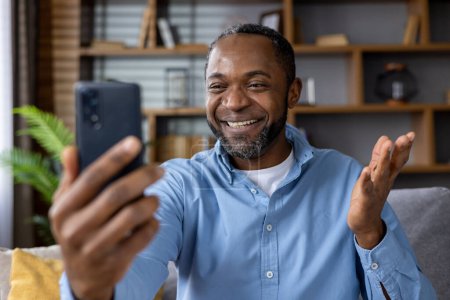Photo for Senior man sitting on sofa in living room, African-American man holding phone in hands, chatting using video call app, smiling joyfully at interlocutor. - Royalty Free Image
