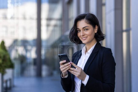 Close-up portrait of a young business woman standing near an office building, holding a phone and smiling at the camera.
