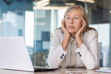 Sad unsatisfied senior woman at workplace depressed, mature business woman unhappy with achievement results at workplace unhappy sitting at table with laptop, boss thinking about problems.