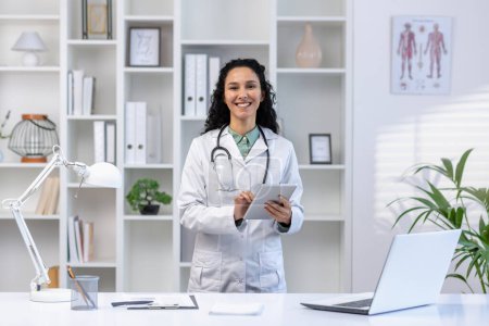 Porträt einer Ärztin im Büro der Klinik, die lächelnd mit einem Tablet-Computer in der Hand am Schreibtisch steht und Patienten aus der Ferne berät.