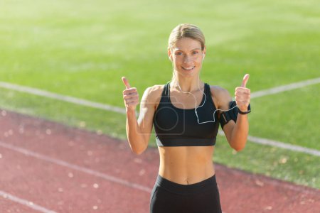 Photo for Athletic woman in sports attire giving a double thumbs up signal on a sunny track field, embodying fitness achievement and positivity. - Royalty Free Image