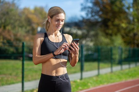 Photo for A fit female athlete in athletic wear selects music on her phone before a run in a sunny park setting. - Royalty Free Image