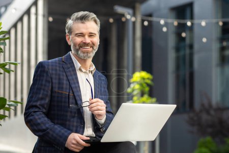Photo for Confident mature businessman with a laptop, smiling outside a modern office building, exuding leadership and success. - Royalty Free Image