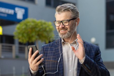 Photo for Mature, well-dressed senior businessman waving while using his phone, standing outdoors near an office building. - Royalty Free Image