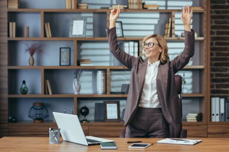 Foto de Mujer profesional madura alegre con los brazos levantados en triunfo en una configuración elegante de la oficina en el hogar, exudando confianza y éxito. - Imagen libre de derechos