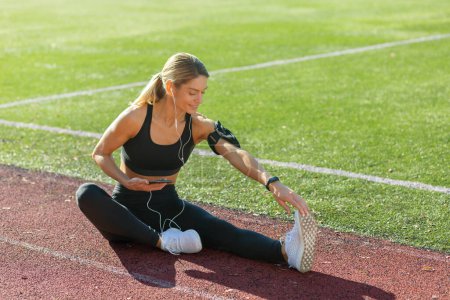 Téléchargez les photos : Une jeune athlète féminine étirant sa jambe sur un terrain d'athlétisme, profitant du soleil du matin avec écouteurs et smartwatch. - en image libre de droit