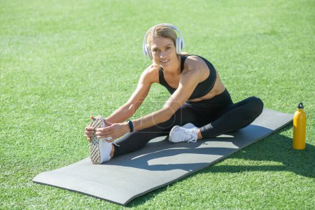 Foto de Ajuste mujer en ropa deportiva disfrutando de la música mientras se estira en una esterilla de yoga. Estilo de vida saludable y rutina de fitness al aire libre capturado. - Imagen libre de derechos