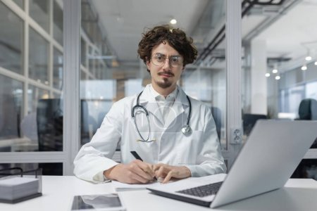 Téléchargez les photos : Un médecin en manteau blanc et stéthoscope est vu assis à un bureau, se concentrant sur son ordinateur portable. Homme rapport écrit sérieux et réfléchi - en image libre de droit