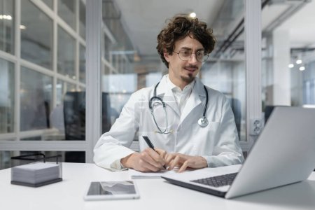 Téléchargez les photos : Un médecin masculin dévoué en manteau blanc et stéthoscope travaillant à son bureau à l'aide d'un ordinateur portable. Homme sourire heureux rapport d'écriture - en image libre de droit